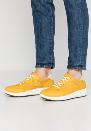 Personne portant un jean bleu et des baskets jaunes vives avec des lacets blancs, debout sur un sol gris clair devant un fond uni.