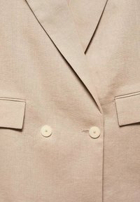 Blazer en lin beige à double boutonnage, avec des revers crantés et deux boutons. Comprend deux poches à rabat. Texture lisse.