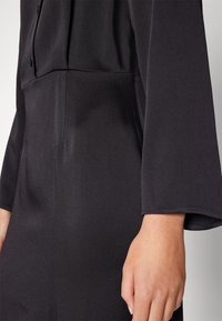 Svart klänning i tyg med slät textur, trekvartsärmar och figurnära midja med knäppningar på framsidan.