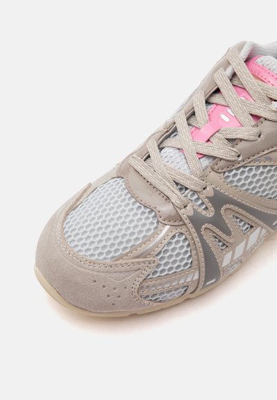 Gros plan sur une basket avec un bout en daim beige, une tige en mesh gris, des empiècements taupe, des lacets beiges et une touche rose sur la languette.