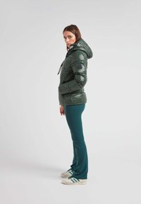 Chaqueta acolchada verde con un acabado brillante, que presenta una capucha, detalles en negro y cordones blancos. Combinada con pantalones verdes y zapatillas.