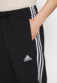 Mustat urheilushortsit valkoisilla sivuraidoilla, joustavalla vyötärönauhalla ja kiristysnarulla, jossa on kirjailtu valkoinen Adidas-logo.