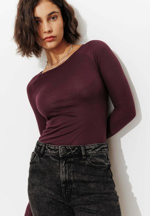 Femme aux cheveux courts et ondulés foncés, portant un haut bordeaux moulant à manches longues et un jean noir taille haute sur fond blanc.