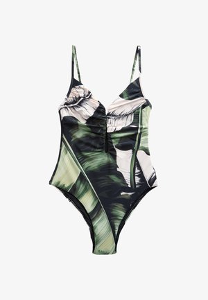 Femme aux longs cheveux foncés portant un maillot de bain une pièce avec des motifs de feuilles vertes et blanches, debout à côté d'un mur en pierre texturé.