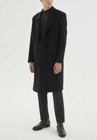 Črn volneni plašč z dolgim, krojenim dizajnom, velikimi ovratniki in stranskimi žepi. Kombiniran s temno sivimi hlačami in rjavimi elegantnimi škornji.