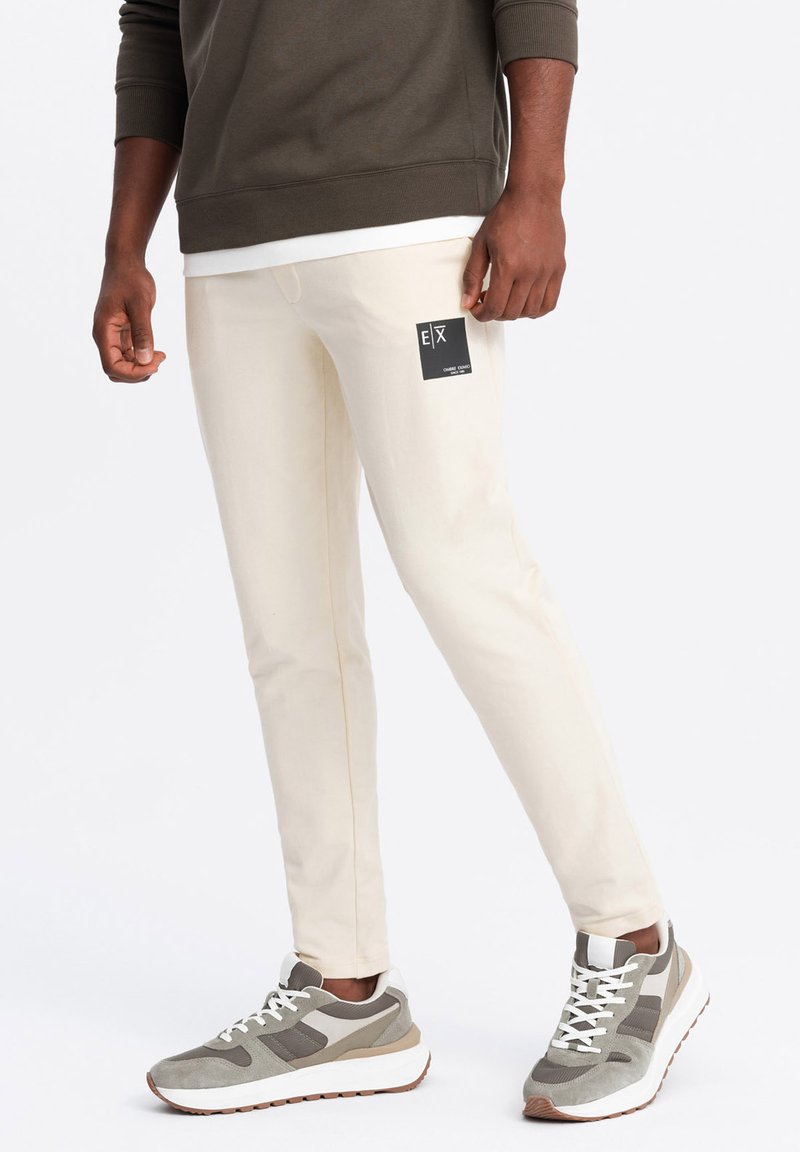 Beige, slim-fit broek met een merklabel in het zwart, gecombineerd met grijze en witte sneakers met een gestructureerd bovenwerk en een dikke witte zool.