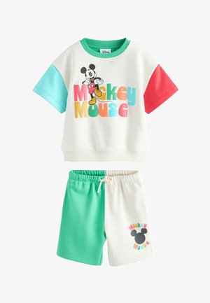 Fiatal fiú, aki színes Mickey egér mintás pólót visel piros, sárga, zöld és türkiz ujjakkal, enyhén felfelé néz egy semleges háttér előtt.