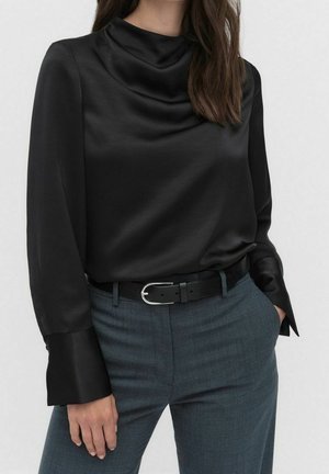 Kvinde iført en sort satinbluse med lange ærmer og en rynket halsudskæring, stukket ned i blå skræddersyede bukser med sort bælte.