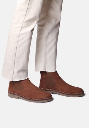 Braune Wildleder Chelsea-Stiefel, getragen mit leicht ausgefransten, off-white Hosen, vor einem hellen, einfarbigen Hintergrund gezeigt.