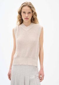 Beige ärmlös stickad tröja med V-ringning, med texturerad tyg och en avslappnad passform, ihop med en ljusgrå plisserad kjol.
