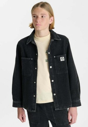 Junge Person mit schulterlangen blonden Haaren, die eine schwarze Cordjacke über einem beigen Hemd vor einem einfarbigen weißen Hintergrund trägt.