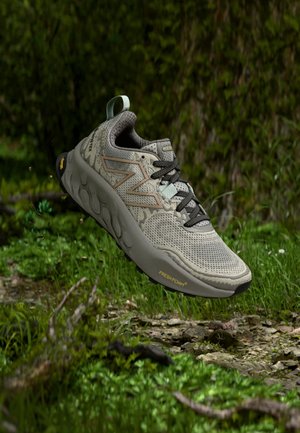 Hellgrauer New Balance Trailrunning-Schuh mit schwarzen Schnürsenkeln und Vibram-Sohle, schwebend über moosigem Waldboden mit grünem Hintergrund.