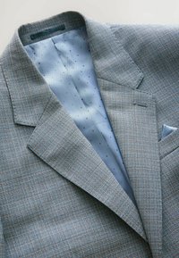 Lys grå blazer med et struktureret vævet stof, udstyret med en blå prikket foring og skarpe klapper. Inkluderer en brystlomme med en matchende detalje.