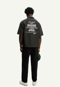 Camisetta nera a maniche corte con testo bianco sul retro. Vestibilità ampia e colletto piatto, abbinata a pantaloni neri e sneakers bianche.