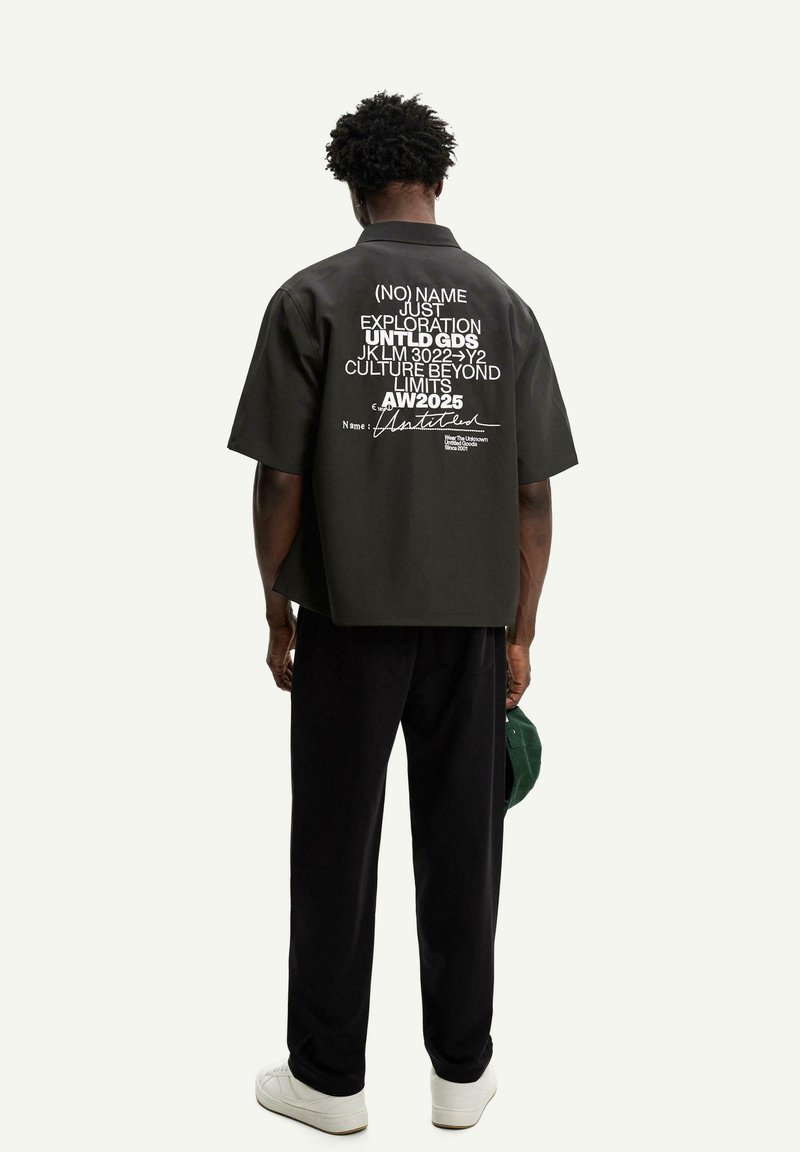 Camisetta nera a maniche corte con testo bianco sul retro. Vestibilità ampia e colletto piatto, abbinata a pantaloni neri e sneakers bianche.