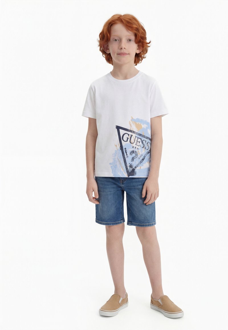 Κοκκινομάλλης αγόρι που στέκεται, φοράει λευκό T-shirt με γραφικό σχέδιο, τζιν σορτς και ανοιχτό καφέ παπούτσια χωρίς κορδόνια, μπροστά σε λευκό φόντο.