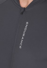 Haut zippé de sport gris foncé fabriqué en tissu lisse et léger. Présente une fermeture éclair centrale et un marquage "ENDURANCE" en blanc.