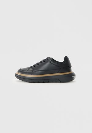 Svart lädersneaker med rund tå, platta snören och en tjock, texturerad gummisula. Har en guldig detalj runt mellansulan.
