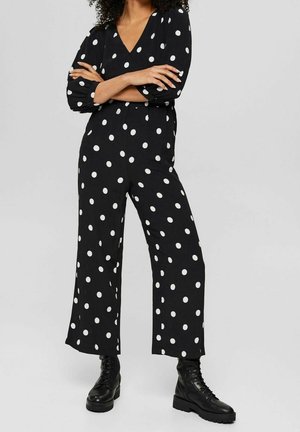 Zwarte jumpsuit met witte stippen, V-hals, driekwartmouwen, losse pasvorm, brede pijpen, gecombineerd met zwarte veterlaarzen.