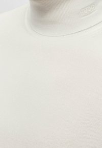 Weißes Rollkragenoberteil aus geripptem Strickmaterial, mit schmaler Passform und dezentem Branding am Kragen. Glatte Textur, keine Muster.