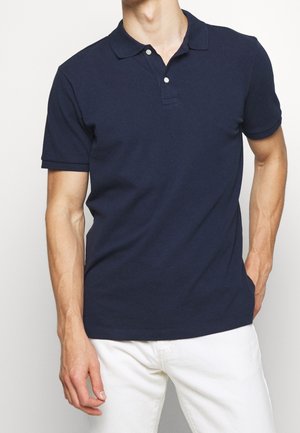 Homme portant un polo bleu marine à manches courtes avec deux boutons, associé à un pantalon blanc, debout devant un fond uni.
