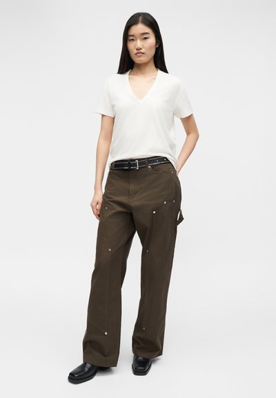 Γυναίκα με μακριά σκούρα μαλλιά που φοράει λευκό κοντομάνικο μπλουζάκι με V λαιμόκοψη, καφέ παντελόνι με φαρδύ πόδι και μαύρη ζώνη, καθώς και μαύρα παπούτσια, στεκόμενη με το ένα χέρι στην τσέπη.