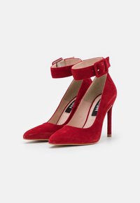 Escarpins rouges en suède à talon haut avec un bout pointu et une bride à la cheville. Dotés d'une fermeture à boucle et d'un talon stiletto fin. Doublure intérieure lisse.