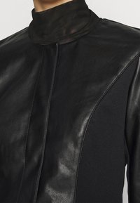 Czarna bluzka z wysokim kołnierzem, wykonana z połączenia skóry i teksturowanej tkaniny, z wyraźnymi szwami i elegancką sylwetką.