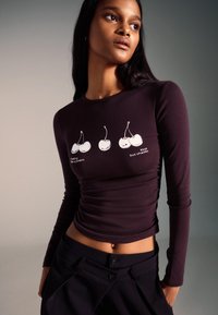 Tmavofialový dlhý rukáv s priliehavým strihom, s grafikou čerešne a textom. Mäkká látka s jemným nariasením pozdĺž strán.