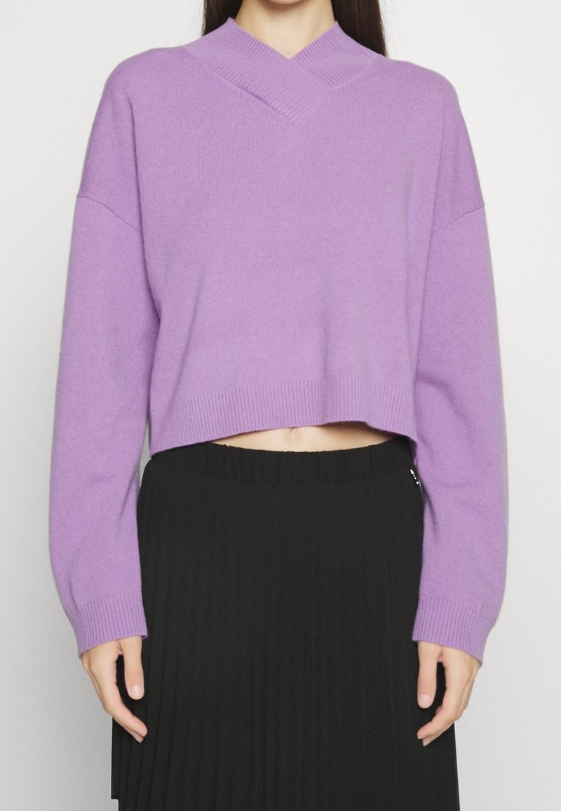 Femme portant un pull court en tricot lavande à col en V et une jupe plissée noire taille haute, debout devant un fond uni.
