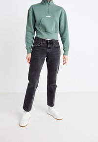 Felpa verde con zip, tasca frontale e logo Levi's, abbinata a jeans neri a vita alta e sneakers bianche.