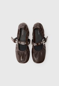 Ballerine in pelle marrone con punta tonda, bordi elasticizzati e una cintura decorativa con fibbia e dettagli metallici. Texture liscia.