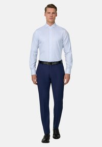 Mann trägt hellblaues Hemd, dunkelblaue Anzughose, schwarzen Gürtel und schwarze Lederschuhe, steht vor einem einfarbigen weißen Hintergrund.