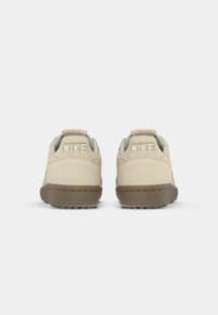 Kreminiai Nike bateliai su zomšiniais viršutiniais, minkštu apykakle ir kontrastingu rudos gumos padu. Iškilmingas logotipas ant kulno.