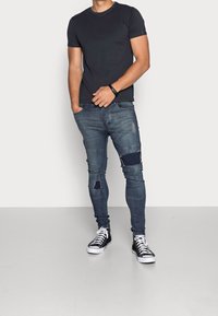 Man som bär svart t-shirt, slitna tight jeans med lappar, svarta högskaftade sneakers, står med en hand i fickan framför en enkel bakgrund.