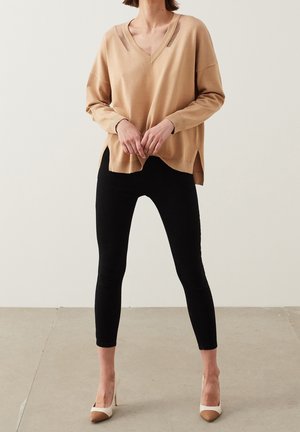 Pull beige en tricot avec un col en V et des fentes latérales, associé à des leggings noirs et des escarpins beiges à bout pointu. Texture lisse et coupe décontractée.