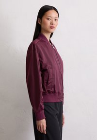 Young woman with long black hair stands sideways wearing a zipped maroon bomber jacket and black pants against a plain wall.