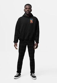 Černoch stojící, oblečený v černé mikině s barevným portrétem Fridy Kahlo na hrudi a černých kalhotách s bílými mřížkovanými liniemi, černých teniskách.