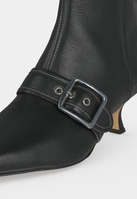Gros plan d'une botte noire en cuir pour cheville avec un bout pointu, une bride à boucle argentée et un talon bas incliné sur fond clair.