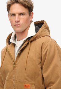 Sudadera de algodón marrón con cierre de cremallera al frente, capucha con cordón, dos bolsillos delanteros y una etiqueta naranja. Presenta una capa interna gris y un cuello blanco.