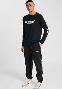 Schwarzes Langarm-T-Shirt mit weißem "hummel"-Logo, kombiniert mit schwarzen Jogginghosen mit weißem Seitenchevron-Design und einem Logo. Graue Sneakers.