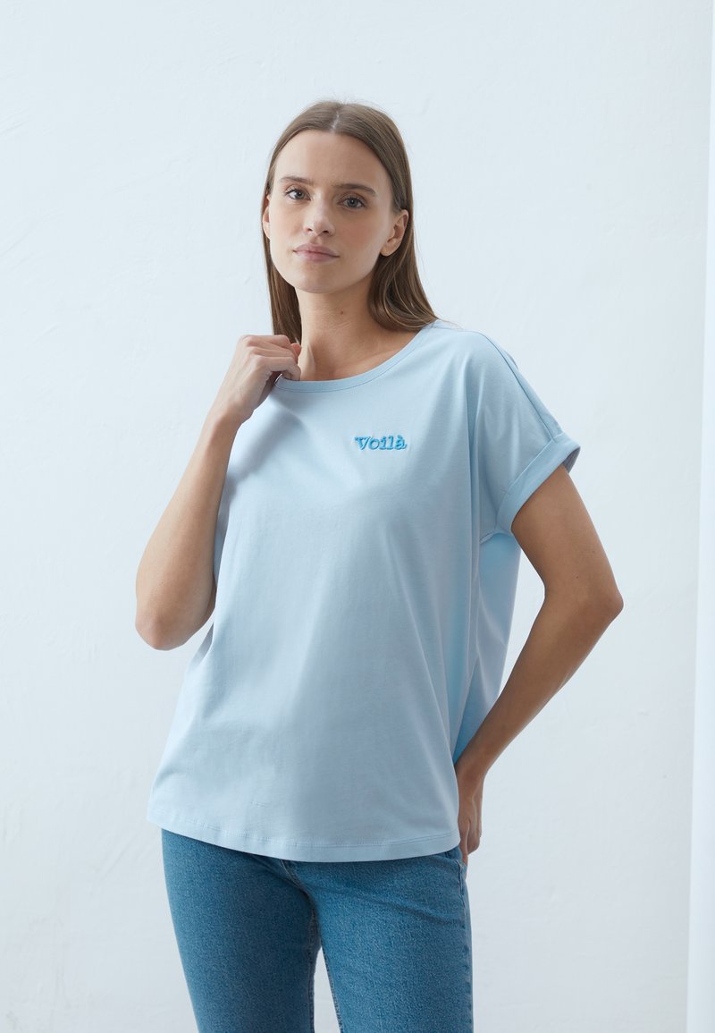 Lysblå bomulds t-shirt med rund halsudskæring og oprullede ærmer, der har den broderede tekst "Voilà" på venstre bryst.