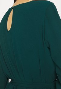 Zielona, teksturowana sukienka z wycięciem na plecach, długimi rękawami i wiązanym paskiem w talii, który podkreśla sylwetkę.