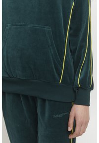 Tuta in velluto di un verde intenso con strisce gialle ai lati. Presenta una tasca frontale e polsini a coste, texture morbida e dettaglio del logo ricamato.