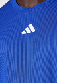 Modra rebrasta športna majica z okroglo ovratnikom in belim logotipom Adidas s tremi diagonalnimi črtami na zgornji levi strani.