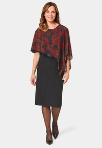 Robe noire avec une superposition fluide et ornée de motifs rouges et noirs. La superposition présente des motifs floraux et un bord en forme de scallop. Escarpins noirs portés.
