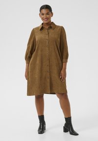 Woman standing, wearing a brown button-up corduroy dress with three-quarter sleeves and black heeled ankle boots, smiling against a plain background.