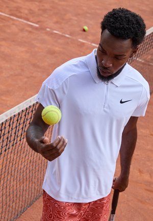 Weißes Nike-Polo mit Reißverschlusskragen, strukturierten Schultern und roten gemusterten Shorts. Spieler balanciert einen gelben Tennisball über einem Sandplatz.