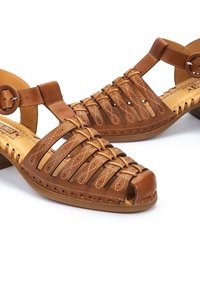 Sandales en cuir marron avec un bout ouvert, design tressé et un petit talon. Elles disposent d'une boucle ronde et de coutures décoratives le long des bords.