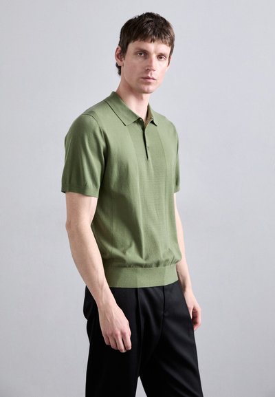 Polo-shirt vert à manches courtes avec un motif texturé, un col polo et une coupe ajustée, porté avec un pantalon noir.